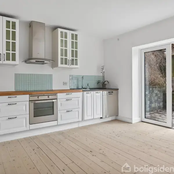 Køkken med hvide skabe og træbord, integreret ovn og emhætte, åbner ud til terrasse gennem glasdør. Rummets lyse vægge og gulv giver et åbent, lyst indtryk.