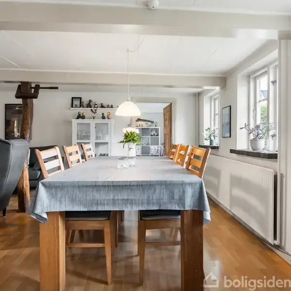 Langt spisebord med blå dug og blomsterdekoration står i spisestue med træmøbler og sofa. Store vinduer på højre væg giver naturligt lys, mens trægulv skaber en varm atmosfære.