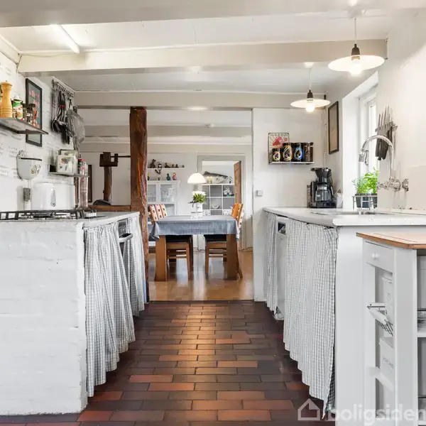 Et lyst køkken med hvide skabe og træelementer, hvor kaffemaskine og planter står på bordet. Baggrunden viser en spisestue med et bord og stole. Lyst og hjemligt interiør.