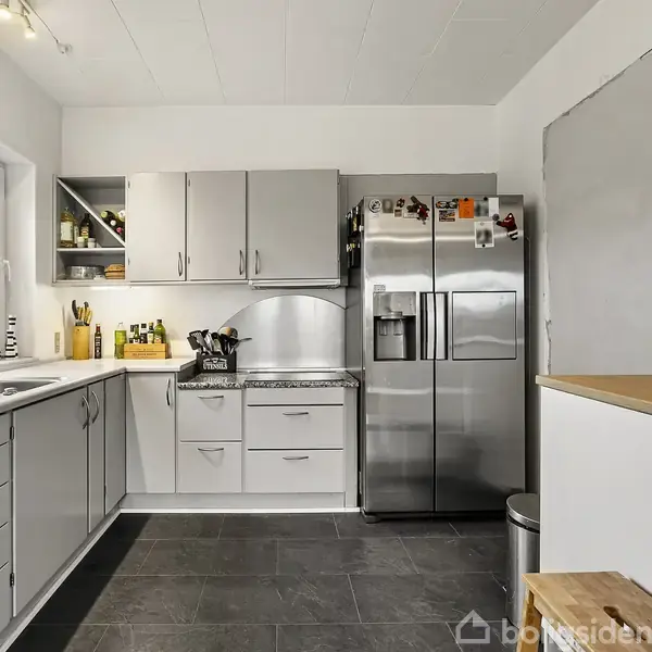 Et moderne køkken med grå skabe og stål køleskab. Opvaskemaskine og vask ved et vindue til venstre. Kaffemaskine og krydderier på træbord til højre. Fliser på gulvet.