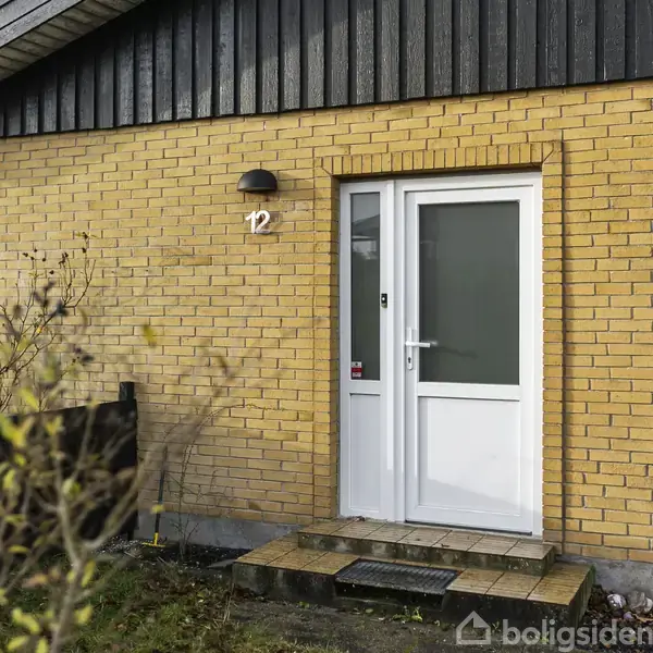 Dør står lukket på en gul murstensbygning. "12" er skrevet ved siden af døren. Et sort hegn og planter omkranser indgangen i en havebeliggenhed.