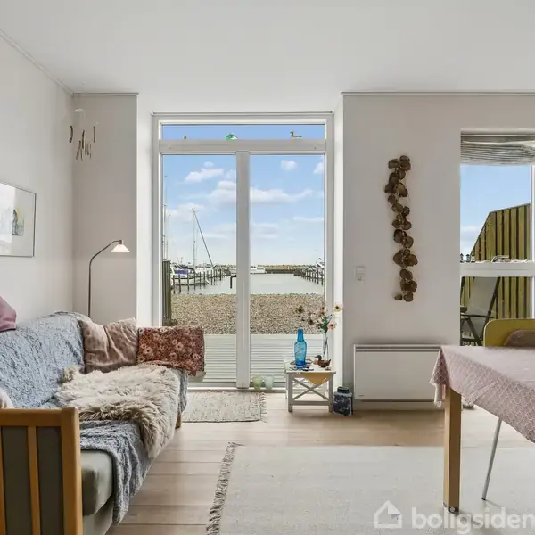 Stue med sofa dækket af tæpper, bordlampe, og spisebord med dug. Gennem glasdøre ses en altan med udsigt til havn og himmel. Dekorationer og billeder på væggene.