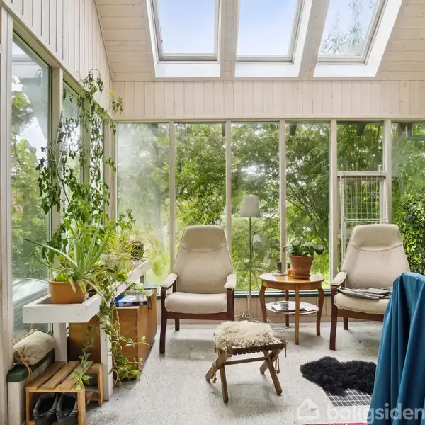 To komfortable stole står ved et lille bord i en lys udestue med store vinduer og loftvinduer. Grønne planter omkranser rummet, og der hænger farvede dekorative tallerkener på væggen.