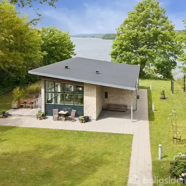 Et moderne hus med fladt tag står i en grøn have tæt på en sø. Der er haveborde og stole på terrassen, omgivet af frodig vegetation.
