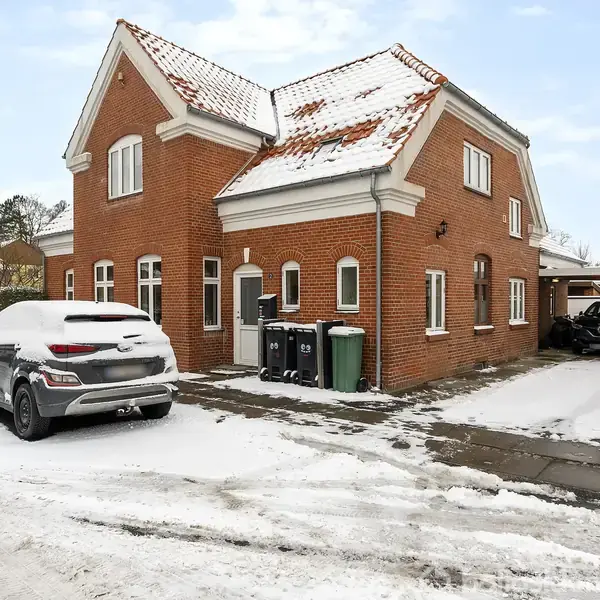 Rødt murstenshus med sne på taget står på en snedækket vej. En bil er parkeret foran, og flere skraldespande er placeret langs husets væg.