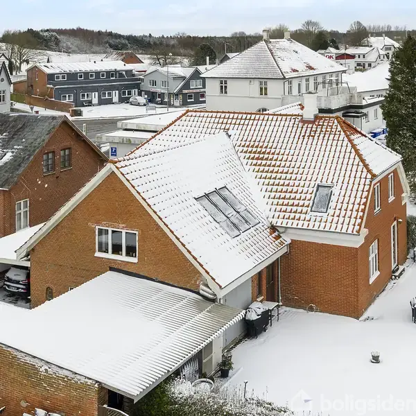 Rødstenshus med snedækket tag står stille blandt andre sneklædte bygninger i et beboelseskvarter. En gårdhave med hvide møbler ses ved siden af. Sne dækker omgivelserne.