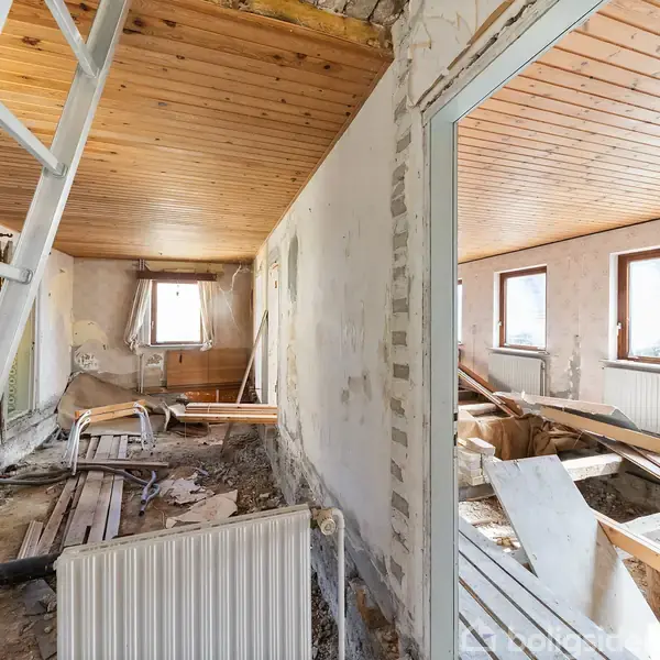 Et rum under renovering med ødelagte vægge og gulvbrædder. En stige står op ad væggen. Der er synlige vinduer, lys træloft, og rod af byggeaffald.