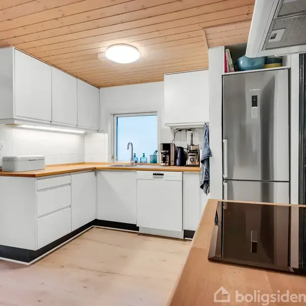 Et moderne køkken med hvide skabe. Køleskabet står til højre, over komfuret ses en emhætte. Træloft og belysning oplyser rummet. Der er ingen personer til stede.