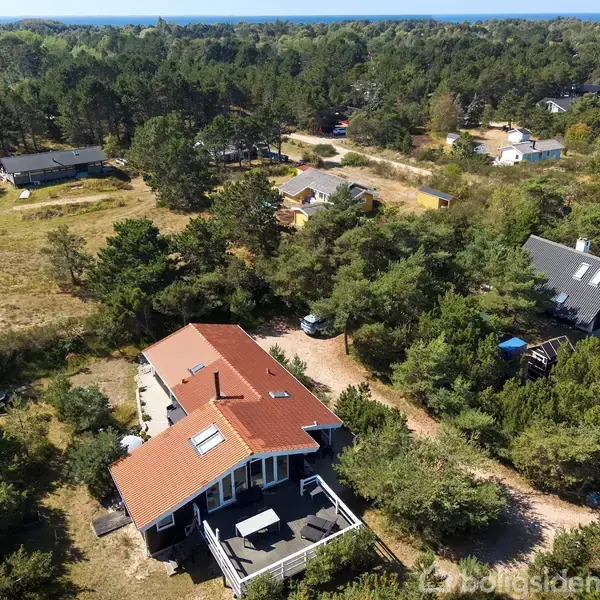 Hus med rød tag og stor terrasse, omgivet af træer og andre huse i en skovklædt, naturskøn landsby. Udsigt over det frodige landskab og nærliggende huse.
