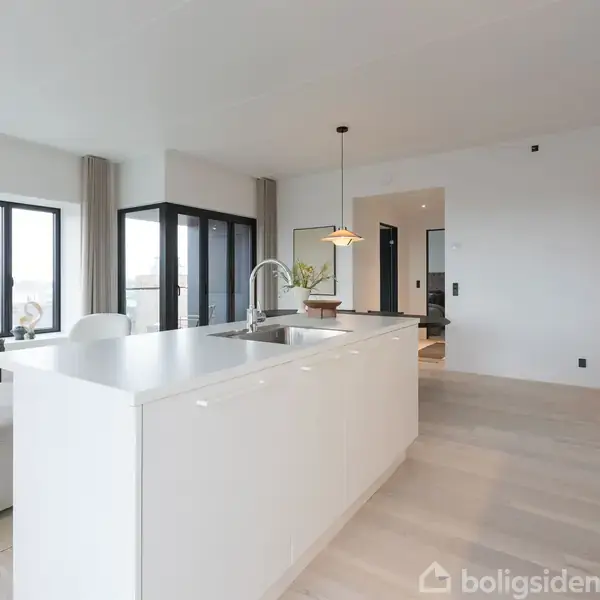 Hvidt køkkenø med indbygget vask står centralt i et moderne, lyst køkken. Store vinduer lader masser af naturligt lys strømme ind i rummet med lys trægulv og minimalistisk indretning.
