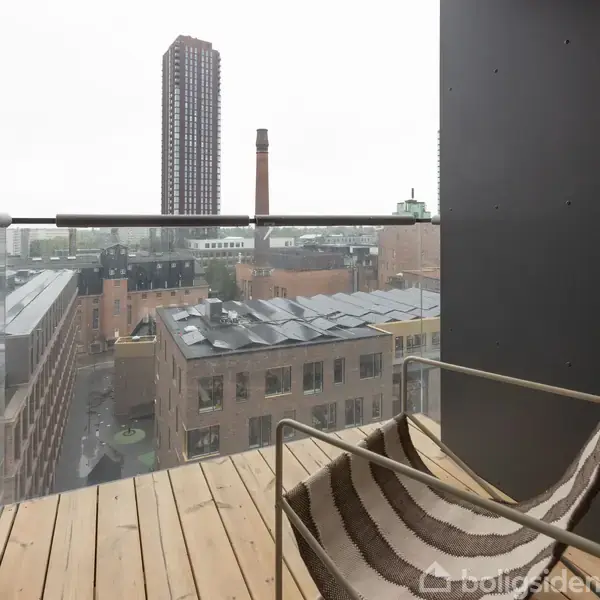 En stol står på en lille altan med trægulv. Den ser ud over en by med høje bygninger, herunder en skorstensbygning og en høj boligblok i baggrunden.