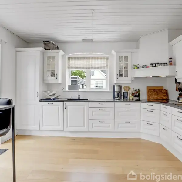 Et lyst, moderne køkken med hvide skabe og stålgreb. Der står køkkenudstyr og krydderiglas på bordpladen. Et vindue giver udsigt til grønne omgivelser. To sorte barstole står ved en bar.