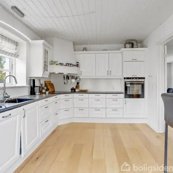 Et moderne køkken med hvide skabe og skuffer, indbygget ovn, mørk bordplade og vask. Rummet har lyse trægulve, vindue til venstre, og et spiseområde med sorte barstole til højre.