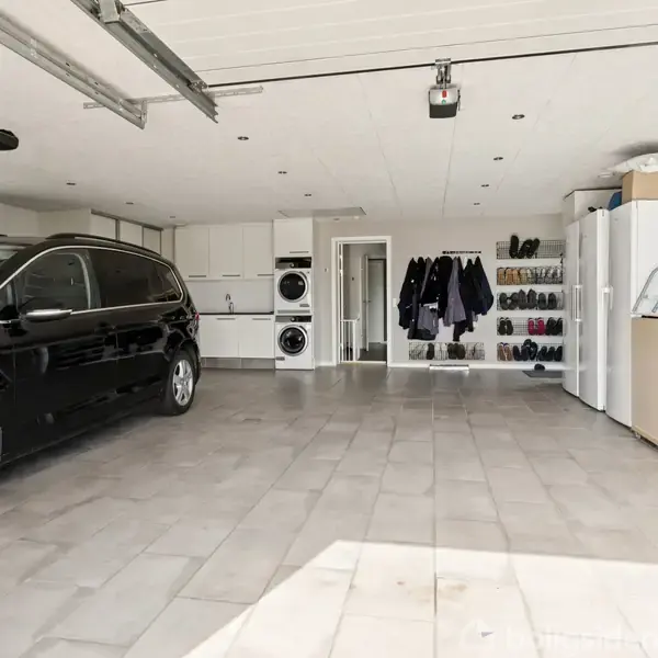 En sort bil holder parkeret i en rummelig garage. Omgivet af vaskemaskine, tørretumbler, jakker hængende og skohylder; flisegulvet er ryddeligt, og et cykelhjul ses til højre.