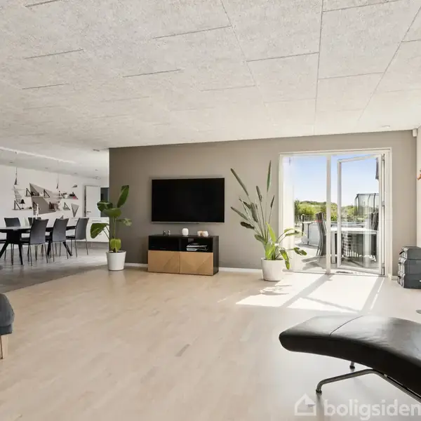 Stue med sofa og lænestol står oplyst af sollys fra en åben altandør. En stor fladskærm hænger på væggen, omgivet af potteplanter. I baggrunden ses et spisebord.