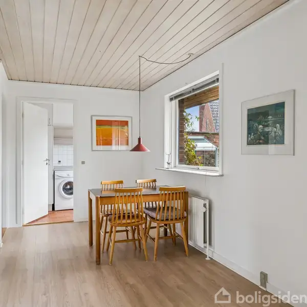 Et spisebord med fire stole står i et lyst køkkenområde. Vinduet viser en have udenfor. I baggrunden er der en dør til et bryggers med en vaskemaskine.