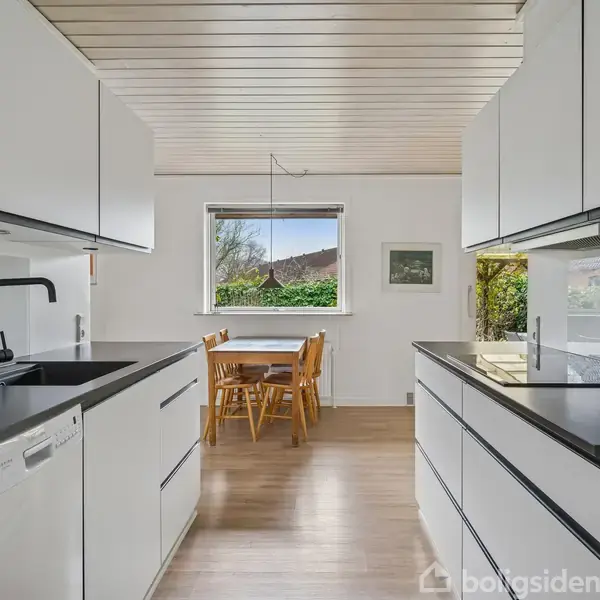 Et moderne køkken med hvide skabe og sort bordplade. Et sort armatur står på vasken. I baggrunden er der et træbord med fire stole ved et vindue ud mod en have.