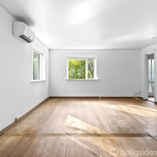 Et tomt rum med trægulv, hvidmalede vægge og to vinduer, som slipper naturligt lys ind. En hvid aircondition-enhed hænger højt oppe på væggen.