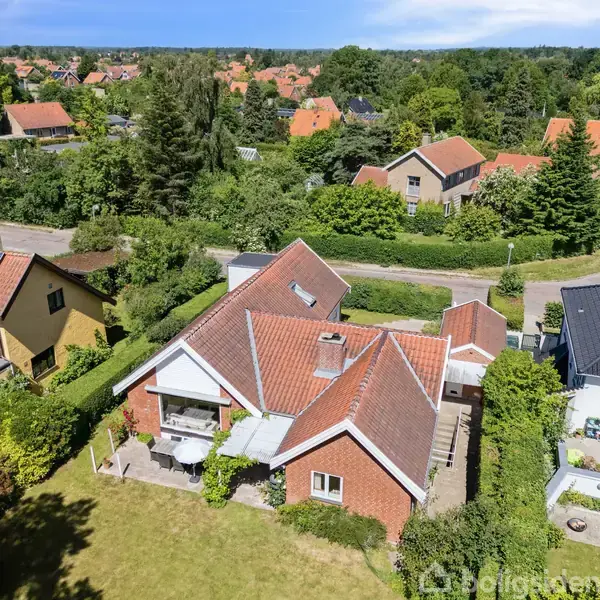 Et rødt murstenshus med et rødt tegltag ses fra oven. En terrasse med bord og stole ses ved siden af huset. Grønt område og flere huse i baggrunden.