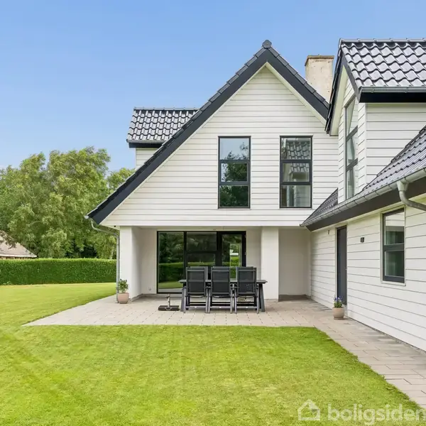 Hvidt, moderne hus med sort tag. Terrasse foran huset har sorte havemøbler, omringet af en velplejet græsplæne og buske. Solrigt vejr med klar blå himmel.