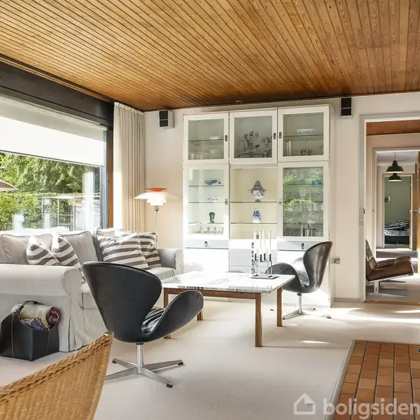 Stue med sofa og moderne stole omkring et sofabord. En reol med glaslåger fyldt med pyntegenstande ses i baggrunden. Stort vindue giver udsigt til grønne omgivelser. Lys træloft.