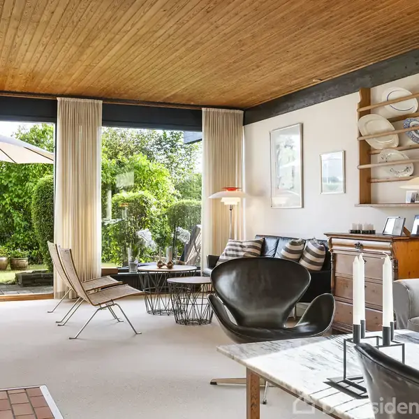 En stue med moderne møbler, herunder en sort designerstol og sofabord. Store vinduer med udsigt til en grøn have skaber en lys atmosfære. Væggene har kunst og hylder med dekorationer.