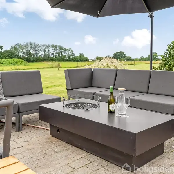 Udendørs terrasse med en grå sofasætning rundt om et sort bord. På bordet står en vinflaske og glas. Baggrunden består af grønne marker og træer under en blå himmel.
