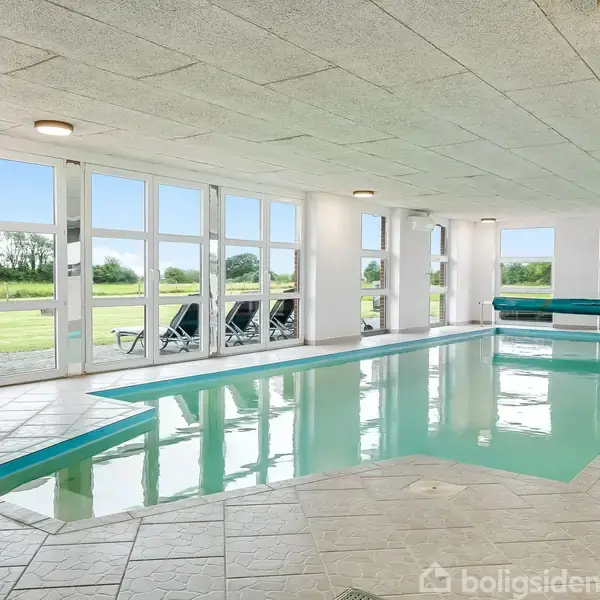 Indendørs swimmingpool med klar blå vandflade, omgivet af hvide fliser. Store vinduespartier afslører en grøn have udenfor med klapstole. Loftet er hvidt med indbyggede lamper.
