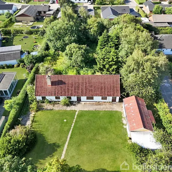 En lang, hvid bygning med rødt tag ligger i en grøn have omgivet af træer og buske, placeret mellem nabo huse. En grusvej fører til en mindre bygning tæt ved.