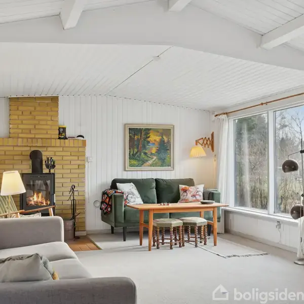 Stue med sofaer og bord, hvor der brænder en ild i pejsen. Store vinduer giver udsigt til natur. Rammerne er hyggelige med lyse vægge og loftsbjælker.