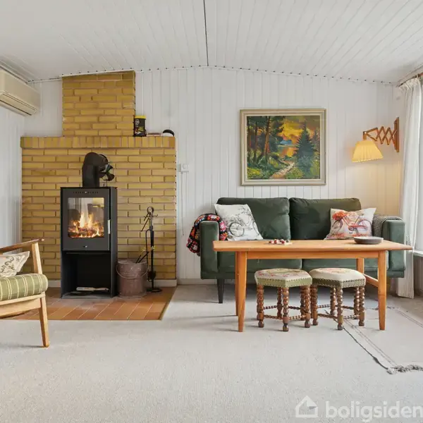 En stue med en brændeovn i midten, omgivet af en grøn sofa, en træbænk og puder. Store vinduer på højre side giver udsigt til udendørsområdet.