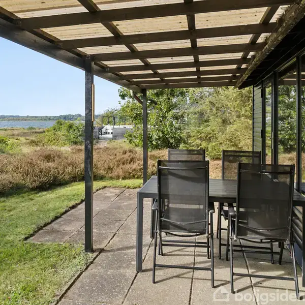 Et sort træhus står midt i en skovklædt naturgrund. Huset har en overdækket terrasse med udendørs bord og stole. Flere træer omgiver stedet, og der er en mindre rød bygning i baggrunden.