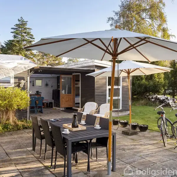 Et bord med stole og parasoller står på en terrasse i en have. Der er cykler ved siden af og forskellige planter og træer omkring, med et hus i baggrunden.