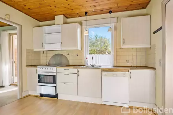 Et køkken med hvide skabe og træbordplader, hvor en komfur og opvaskemaskine er installeret. Træloftet giver varme, og et vindue giver udsigt til en have.