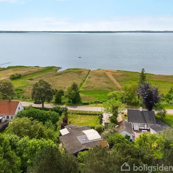Huse står blandt frodige træer nær vandet i en landlig setting. Vejen adskiller bebyggelsen fra de åbne engarealer ved søbredden. Solen skinner klart over landskabet.