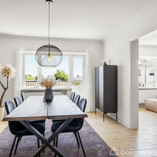 Spisebord med seks mørke stole er placeret på et tæppe, lys hængelampe ovenover. Vegetation i vase og store vinduer i baggrunden. Adgang til lys stue til højre.