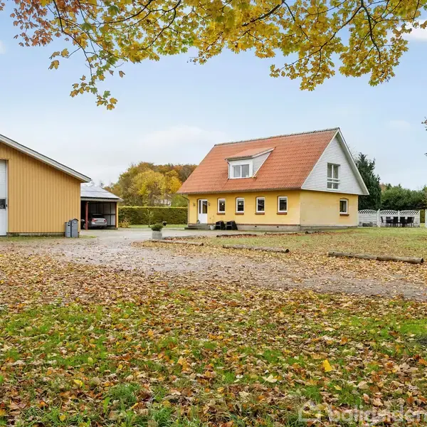 Et gult hus med rød tag ligger ved siden af en stor garagebygning. Omgivet af træer og et tæppe af efterårsblade.