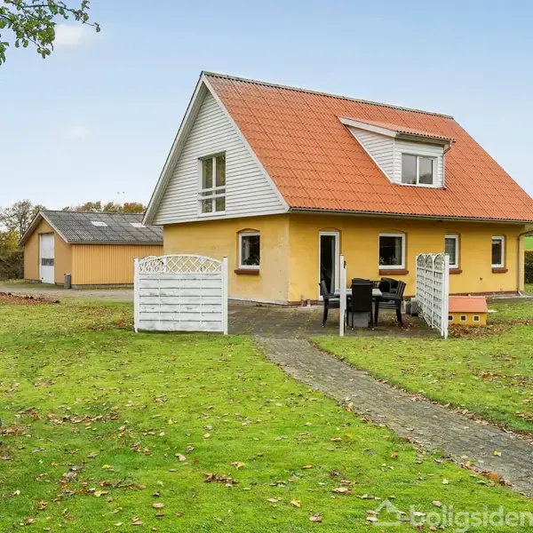 Et gult hus med rødt tag står midt i en grøn græsplæne. En hvid port er åben, og der er en terrasse med havemøbler. Baggrunden viser træer og en garage.