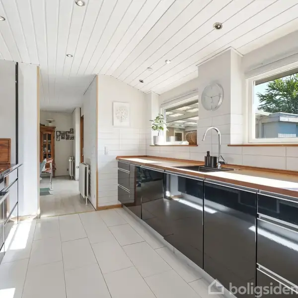Et moderne køkken med sorte skabe og træbordplade, hvide fliser og loft. Solen skinner gennem et stort vindue over vasken, og giver udsigt til en gårdhave.