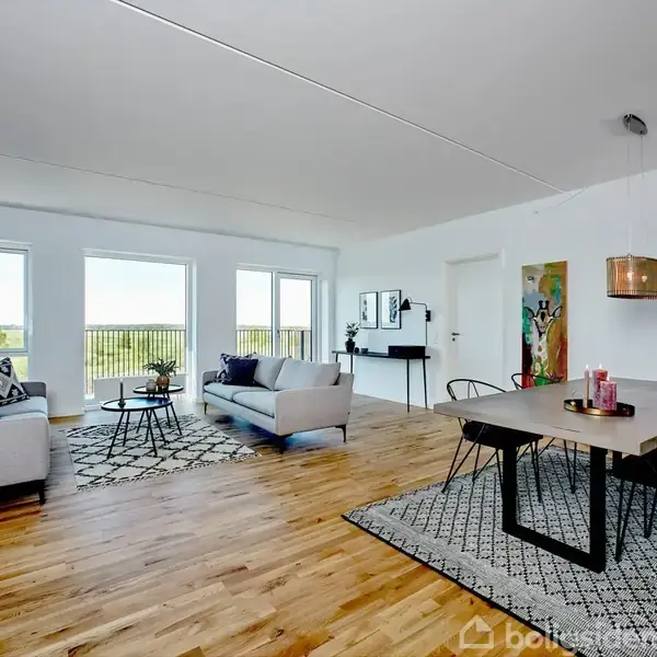 En lys stue med hvide vægge og trægulv indeholder to grå sofaer foran store vinduer, et spisebord med sort stel, og flere kunstværker på væggene.