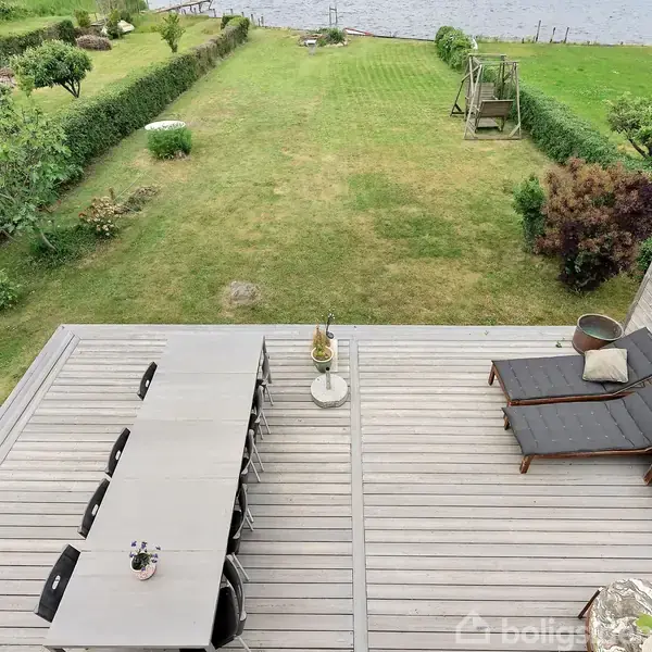 Terrasse med lang spisebord og stole på træplanker, og to liggestole ved siden af. Haven har græsplæne, buske og træer, beliggende ved en sø i baggrunden.