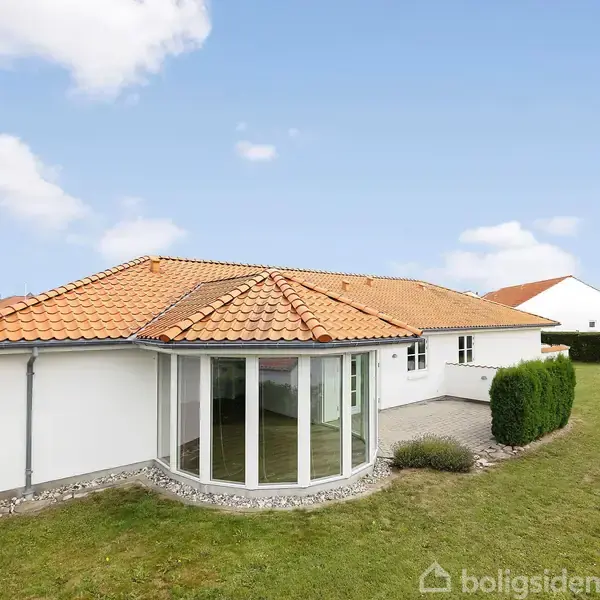 Huset med rødt tegltag har store vinduer og ligger i en velholdt have. Yderligere omgivet af hække og græs, det er placeret i et boligområde med flere lignende huse.
