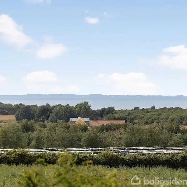 En gul bygning står stille omgivet af grønne marker og buske, med en blå himmel og hav i horisonten. Der er ingen tekst i billedet.