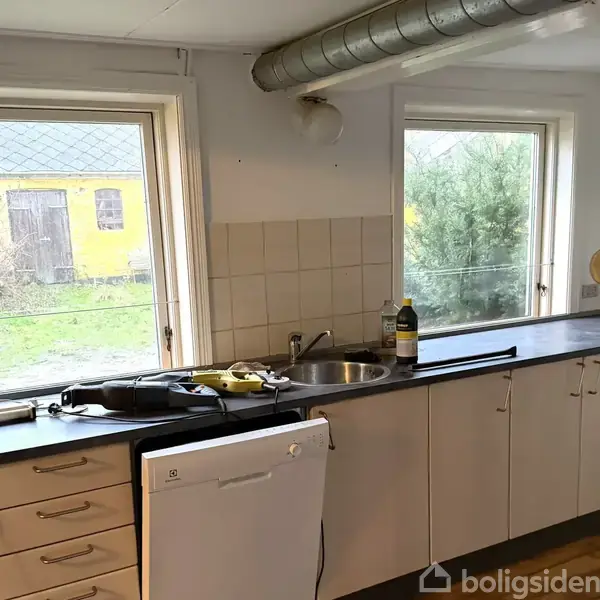 Køkken med hvide skabe og en sort bordplade. En opvaskemaskine står under bordet. Udenfor ses en gul bygning og grønne buske gennem vinduerne.