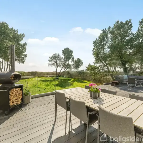 Terrasse med spisebord og stole, hvor en pejs opbevarer brænde. I baggrunden ses grønne træer og have. Sollys skaber en lys og indbydende atmosfære.