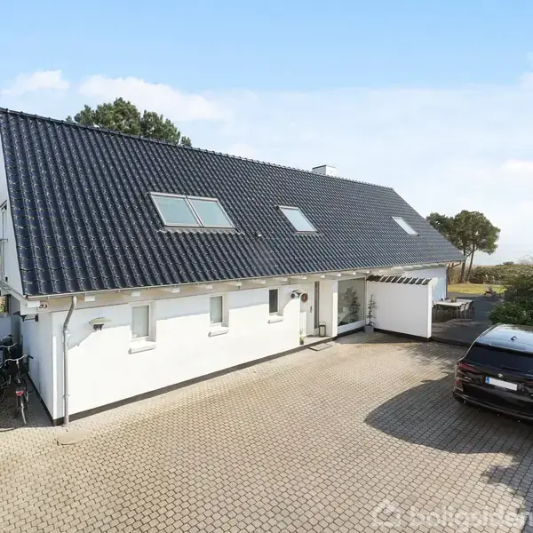 Et hvidt hus med sort tag står på en flisebelagt indkørsel. Cykler er placeret ved siden af huset, og en sort bil holder parkeret. Omgivet af træer og buske.