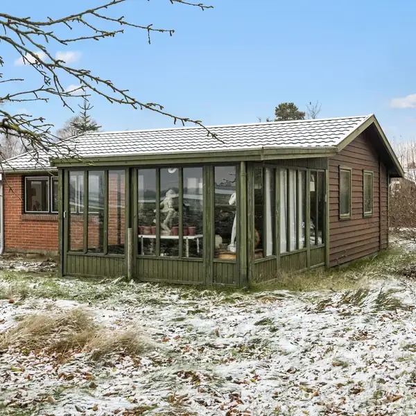 Et lille træhus med glasveranda står i en have dækket af let sne. Skyfri blå himmel og sparsom beplantning omgiver huset.