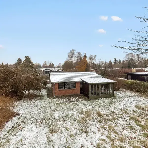 Et lille murstenshus med hvide vinduer står i en have dækket af et let lag sne, omgivet af buske og træer, under en klar, blå himmel.