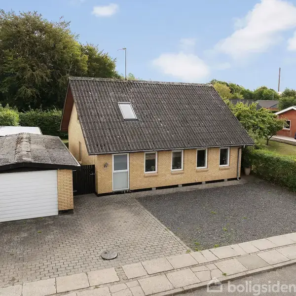 Et gult murstenshus med sort skråtag står stille ved en asfalteret indkørsel og grusgrund, omgivet af grøn hæk og andre boligbygninger i et villakvarter.