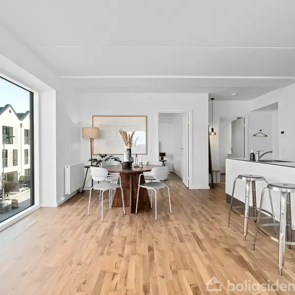 Et lyst og moderne køkken-alrum med trægulv og store vinduer. Rundt spisebord med stole centralt, køkken med barstole til højre, visning mod boligområde udenfor.